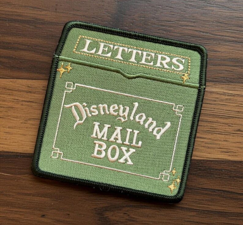May include: Green embroidered patch with the words "LETTERS" and "Disneyland MAIL BOX" in white and gold lettering. The patch has a black border and is rectangular in shape.