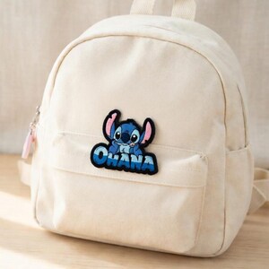 May include: Off-white canvas backpack with a front pocket and top handle. A blue and black patch of the character Stitch with the word "OHANA" is on the front pocket. The backpack has a zipper closure and adjustable straps.