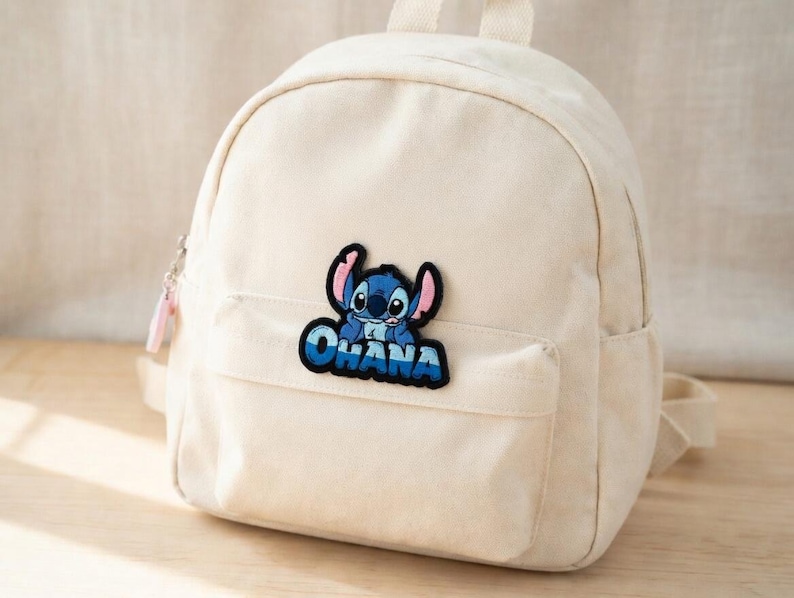May include: Off-white canvas backpack with a front pocket and top handle. A blue and black patch of the character Stitch with the word "OHANA" is on the front pocket. The backpack has a zipper closure and adjustable straps.