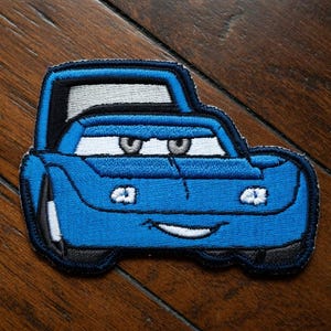 May include: Embroidered blue car patch with black and white detailing. The patch depicts a smiling car with large eyes and a cartoonish design. The patch is likely for clothing or accessories.