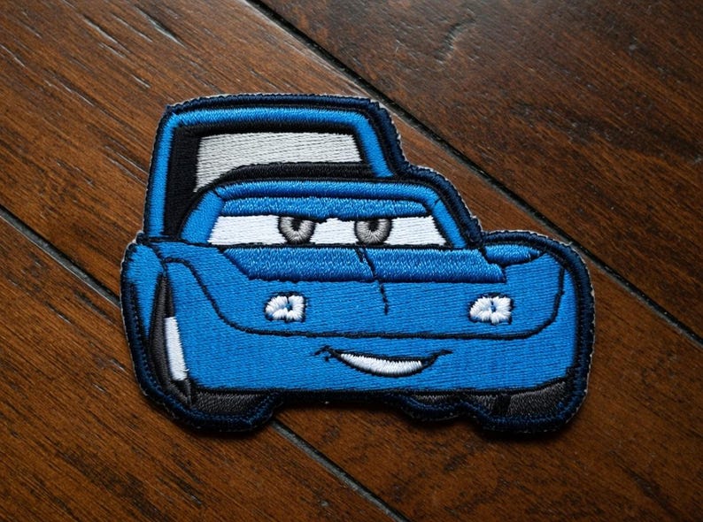 May include: Embroidered blue car patch with black and white detailing. The patch depicts a smiling car with large eyes and a cartoonish design. The patch is likely for clothing or accessories.