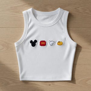 May include: White ribbed crop top featuring embroidered Mickey Mouse-themed motifs. The design includes a black Mickey head, red shorts, a white glove, and yellow shoe. The sleeveless top has a round neckline and is made of a soft, ribbed fabric.