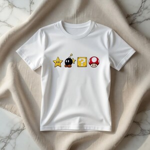 May include: White t-shirt with a cartoon design featuring a yellow star, a black bomb, a yellow question mark block, and a red mushroom. The graphics are from a popular video game.