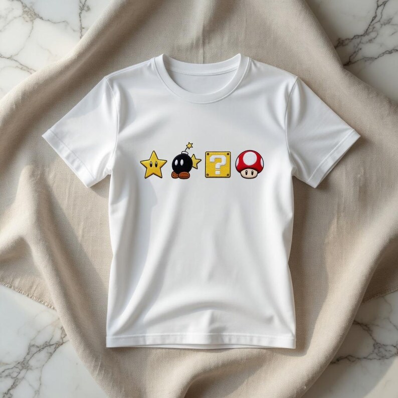 May include: White t-shirt with a cartoon design featuring a yellow star, a black bomb, a yellow question mark block, and a red mushroom. The graphics are from a popular video game.