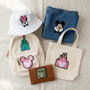 May include: An assortment of Disney-themed accessories, including a white bucket hat with a Minnie Mouse patch, a denim pair of jeans with a Mickey Mouse patch, a canvas tote bag, a backpack, and a brown wallet.