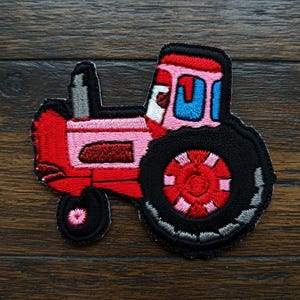 May include: Embroidered patch featuring a cartoon tractor. The tractor is predominantly red and pink, with black tyres and a grey smokestack. The design includes blue window details and a pink and red wheel.