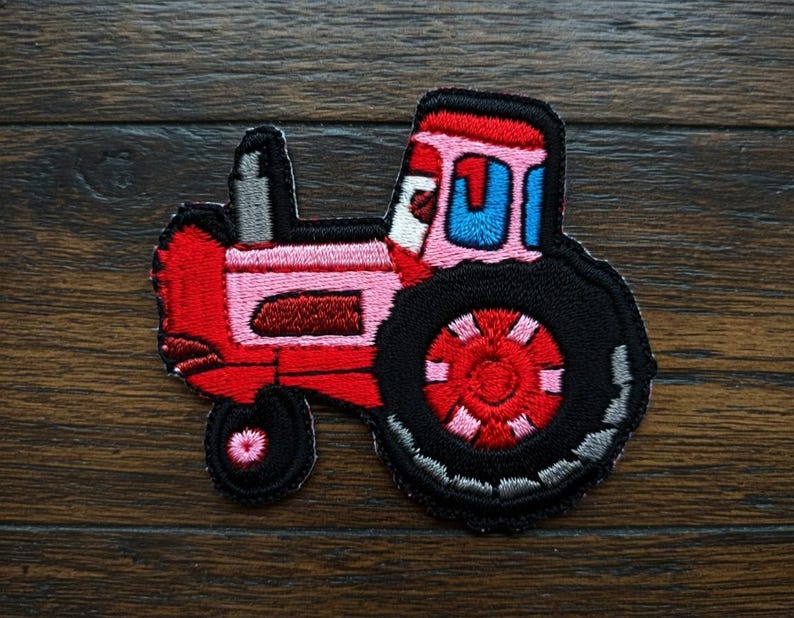 May include: Embroidered patch featuring a cartoon tractor. The tractor is predominantly red and pink, with black tyres and a grey smokestack. The design includes blue window details and a pink and red wheel.