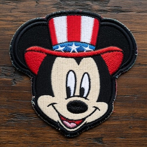 May include: Embroidered patch featuring Mickey Mouse wearing a patriotic Uncle Sam hat with red and white stripes and blue stars. The patch has a black border and a smiling face.