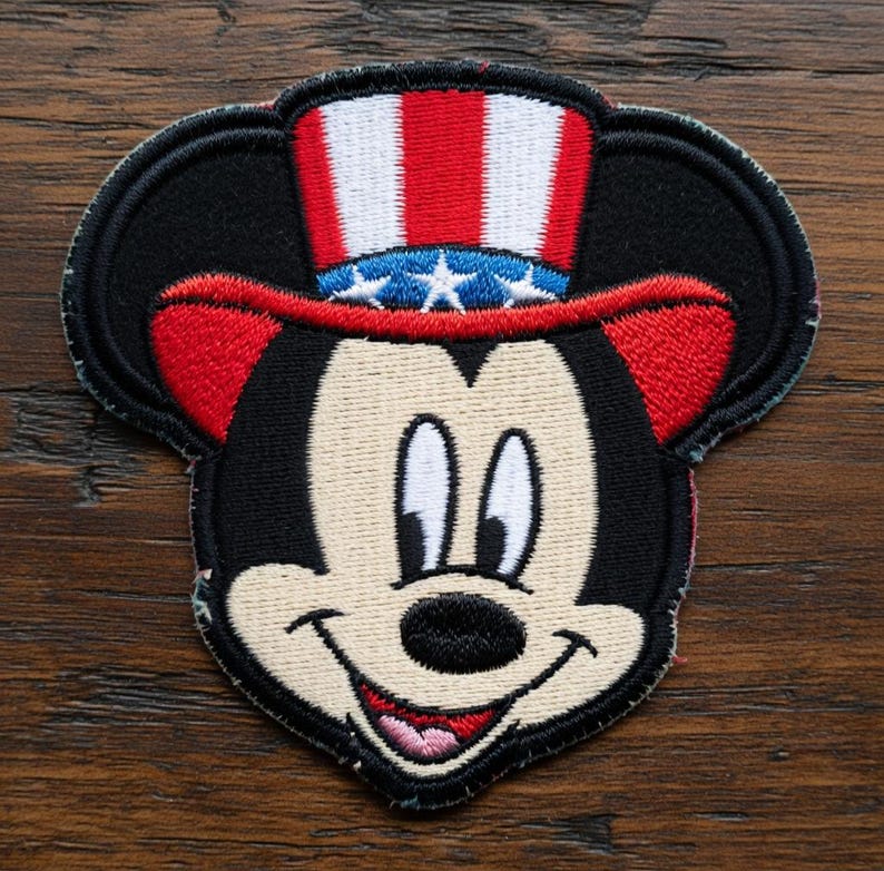 May include: Embroidered patch featuring Mickey Mouse wearing a patriotic Uncle Sam hat with red and white stripes and blue stars. The patch has a black border and a smiling face.