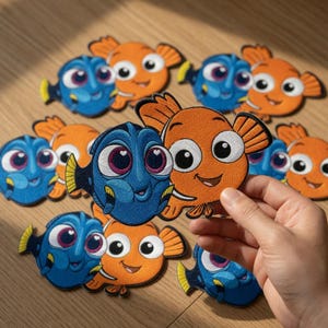 May include: Embroidered patches featuring cartoon fish characters in blue and orange. The patches have detailed facial features and are arranged on a wooden surface. The patches are likely for clothing or accessories.