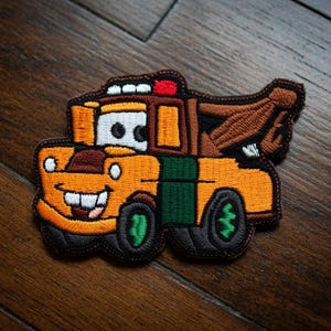 May include: Embroidered patch of a cartoon tow truck with a smiling face. The patch is mainly orange with green and brown details. The tow truck has a red light and black tyres.