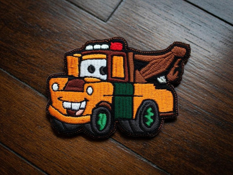 May include: Embroidered patch of a cartoon tow truck with a smiling face. The patch is mainly orange with green and brown details. The tow truck has a red light and black tyres.