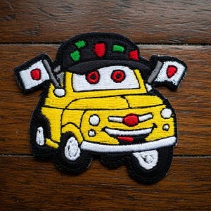 May include: Embroidered patch of a cartoon car with a smiling face, red eyes, and a red nose. The yellow car has black tires and a black hat with red and green accents. Two small flags with a red circle on a white background are attached.