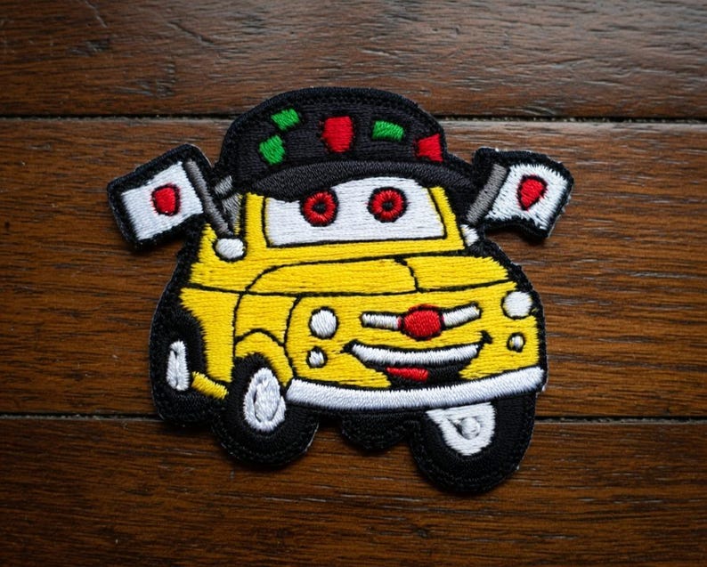 May include: Embroidered patch of a cartoon car with a smiling face, red eyes, and a red nose. The yellow car has black tires and a black hat with red and green accents. Two small flags with a red circle on a white background are attached.