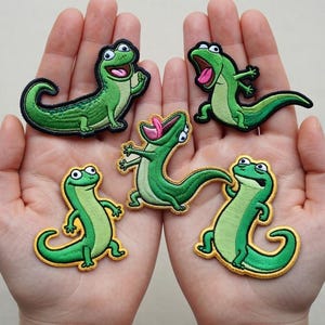 Pode incluir: Cinco patches bordados de lagartos verdes com contornos pretos e bordas amarelas. Cada lagarto tem uma pose diferente, com bocas abertas e olhos expressivos. Os patches são segurados em mãos abertas.