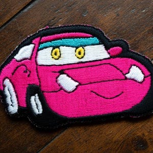 May include: Embroidered patch of a cartoon car in pink, black, and white. The car has yellow eyes, a turquoise visor, and a smiling mouth. The patch is designed for application on clothing or accessories.