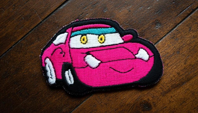 May include: Embroidered patch of a cartoon car in pink, black, and white. The car has yellow eyes, a turquoise visor, and a smiling mouth. The patch is designed for application on clothing or accessories.