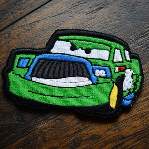 May include: Embroidered patch depicting a cartoon car. The car is predominantly green, with blue, black, white, and yellow details. The patch has a black border and is designed for application on clothing or accessories.