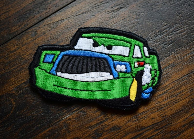 May include: Embroidered patch depicting a cartoon car. The car is predominantly green, with blue, black, white, and yellow details. The patch has a black border and is designed for application on clothing or accessories.