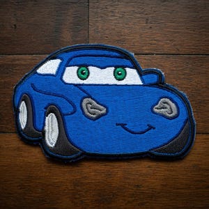May include: A blue embroidered car patch with a smiling face and green eyes. The patch has white accents and black tyres. The car is a cartoon style and is on a wooden surface.