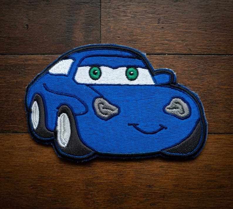 May include: A blue embroidered car patch with a smiling face and green eyes. The patch has white accents and black tyres. The car is a cartoon style and is on a wooden surface.