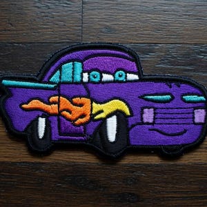May include: Embroidered patch of a cartoon car in purple, teal, orange, and yellow. The car has a flame design and white wheels with black tyres. The patch is outlined in black, suitable for clothing or accessories.