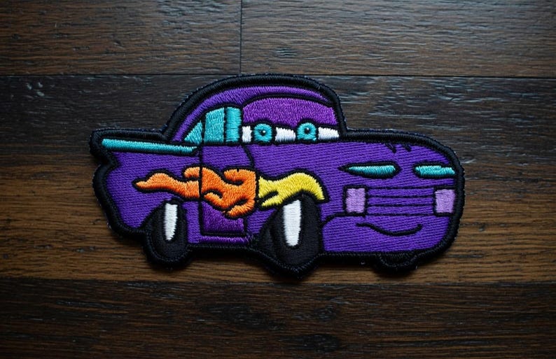May include: Embroidered patch of a cartoon car in purple, teal, orange, and yellow. The car has a flame design and white wheels with black tyres. The patch is outlined in black, suitable for clothing or accessories.