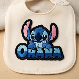 May include: Cream-colored baby bib with a blue and black embroidered image of the character Stitch from Lilo & Stitch. The word "OHANA" is embroidered in blue and black below the character.