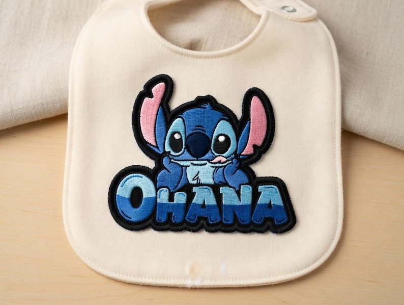 May include: Cream-colored baby bib with a blue and black embroidered image of the character Stitch from Lilo & Stitch. The word "OHANA" is embroidered in blue and black below the character.