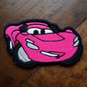 May include: A pink embroidered patch in the shape of a cartoon race car. The car has white and black accents, including the eyes, tyres, and outline. The patch is on a dark wood surface.