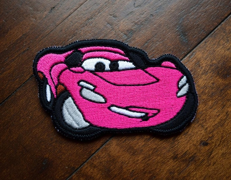 May include: A pink embroidered patch in the shape of a cartoon race car. The car has white and black accents, including the eyes, tyres, and outline. The patch is on a dark wood surface.