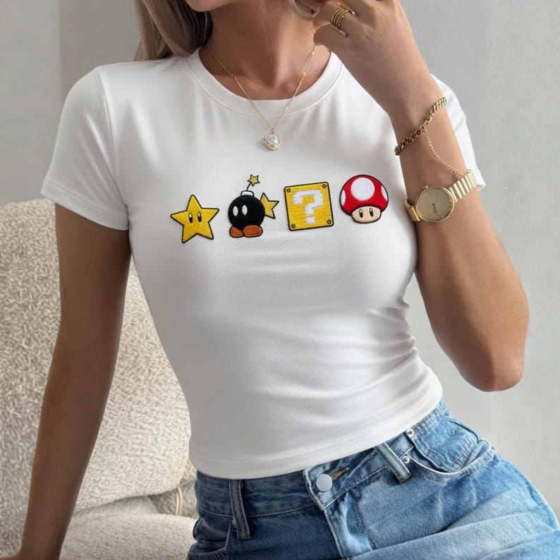 May include: White, short-sleeved t-shirt with embroidered video game graphics. The design includes a yellow star, a bomb, a question mark box, and a red mushroom. The shirt is made of a soft material.