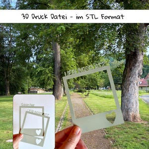 Può includere: Una cornice stampata in 3D verde chiaro a forma di foto quadrata con il testo LIEBLINGSPLATZ in alto e un ritaglio a forma di cuore in basso. La cornice è tenuta in mano davanti a un parco. Il testo in alto nell'immagine recita 3D Druck Datei - im STL Format.