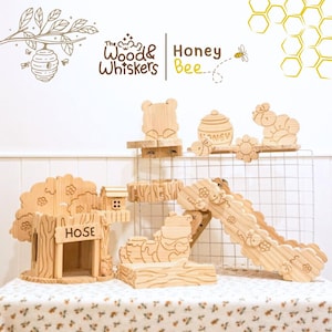 May include: Wooden toy set featuring a bear, honey pot, and other bee-themed shapes. The set includes a treehouse with a "HOSE" sign, a slide, and various platforms. The Wood & Whiskers logo is visible.