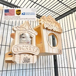 May include: A light-colored chinchilla peeks out from a wooden ticket booth-shaped structure. The booth has the word "TICKETS" carved on it. Next to it is a wooden house with a "POPCORN" sign. The structures are inside a wire cage.
