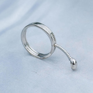 May include: A silver ring with a small, dangling ball. The ring has a simple band design with a small, decorative element. The ring is set against a light blue background.