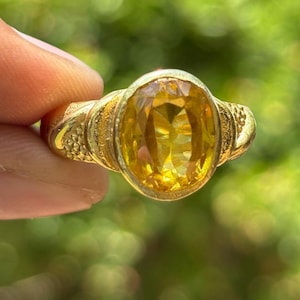 Peut inclure: Une bague en or ornée d'une pierre précieuse jaune ovale. La bague a un anneau et un sertissage texturés, avec un aspect brillant et poli. La pierre précieuse est d'un jaune vif, sertie dans une monture en or détaillée.