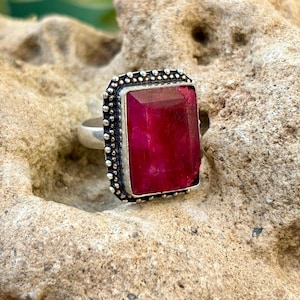 May include: A silver ring with a large, rectangular, deep red gemstone. The gemstone is set in a square frame with a beaded border. The ring is displayed on a textured, light brown surface, with green foliage in the background.