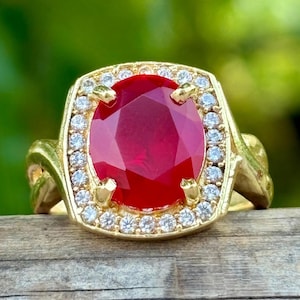 Può includere: Un anello d'oro con una grande pietra preziosa rossa intensa a taglio ovale, circondata da un alone di piccole gemme trasparenti. La fascia dell'anello presenta un design ornato e strutturato. Lo sfondo è verde sfocato.