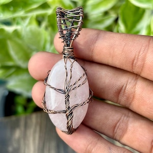 Op de afbeelding: Een rozenkwarts hanger met een boomontwerp van koperdraad. De ovale roze steen is omwikkeld met koperdraad, waardoor een boommotief ontstaat. De hanger hangt aan een gevlochten koperdraad beugel.