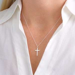 Può includere: Una collana con pendente a forma di croce argentata. La croce è di design semplice, sospesa a una delicata catena. La collana è indossata su una camicia bianca.