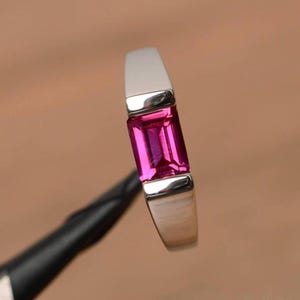 May include: A silver ring with a rectangular, vibrant pink gemstone. The gemstone is set in a polished silver band, creating a modern and elegant design. The ring is held against a blurred background.