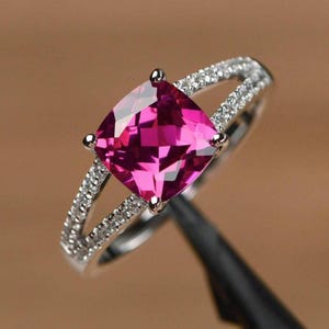 Op de afbeelding: Een zilverkleurige ring met een grote, kussengeslepen, levendige magentakleurige edelsteen in het midden. De ringband is gespleten in twee delicate armen, die beide over de gehele lengte zijn ingelegd met kleine, heldere, fonkelende witte steentjes.