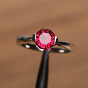Könnte beinhalten: Ein silberner Ring mit einem auffälligen, achteckigen, rubinroten Edelstein. Der Edelstein ist das zentrale Merkmal, eingebettet in ein schlichtes, elegantes Band. Das Design des Rings ist klassisch und zeitlos.