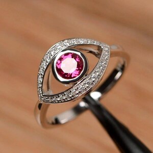 Könnte beinhalten: Ein silberner Ring mit einem zentralen, runden, rosa Edelstein. Der Edelstein ist in einer Lünette gefasst. Das Ringband und die Lünette sind mit kleinen, klaren Edelsteinen besetzt, wodurch ein funkelnder Effekt entsteht. Das Design ähnelt einem Auge.