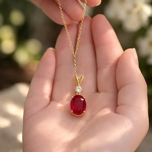 Puede incluir: Un collar de oro con un gran colgante ovalado de piedra preciosa rojo oscuro. El colgante está acentuado con una pequeña piedra preciosa clara y redonda. El collar se sostiene en una mano abierta sobre un fondo desenfocado.