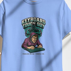 May include: Light blue t-shirt with a graphic featuring the text "KEYBOARD TOUGH GUY" above an illustration of a person typing on a keyboard. The graphic includes a muscular figure in the background. The text "FEARLESS ONLINE - FRAGILE IRL" is below the illustration.