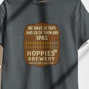 Op de afbeelding: Donkergrijs t-shirt met een bruine opdruk met de tekst "We have 30 taps and 28 of them are IPAs. Hoppies Brewery, Sweetgum Junction." De opdruk bevat illustraties van biertaps.