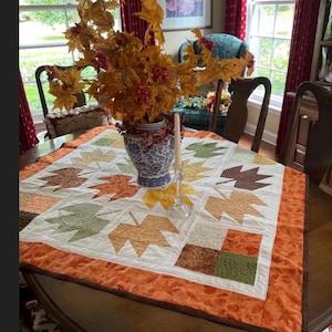 May include: A square quilted table topper with a leaf pattern in shades of orange, yellow, and green. The topper is bordered with an orange fabric. A vase of fall foliage and a candle sit on the table, creating a seasonal display.