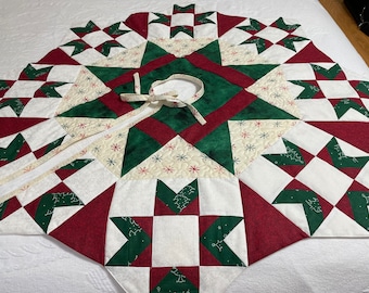May include: A quilted Christmas tree skirt with a star pattern. The skirt features a patchwork design with red, white, and green fabric. The skirt has a cream-colored tie closure and is decorated with snowflake and Christmas tree patterns.